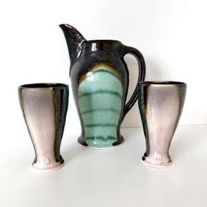 May include: A ceramic pitcher and two matching tumblers featuring a glossy glaze with a central vertical panel of light green transitioning into dark brown and black edges. Each piece has a flared base and a smooth, rounded silhouette.