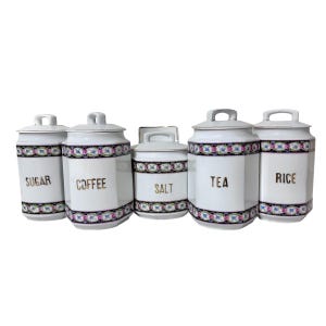 May include: A set of five white ceramic kitchen canisters with lids. Each canister is labeled with gold lettering: Sugar, Coffee, Salt, Tea, and Rice. A decorative band of pink, blue, and green floral pattern encircles each canister.