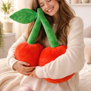 Könnte beinhalten: Ein Plüsch-Kirschkissen mit zwei großen roten Kirschen, grünen Stielen und einem grünen Blatt. Ideal für die Heimdekoration oder als Geschenk. Die Kirschen haben eine leuchtend rote Farbe.