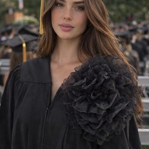 May include: A person wearing a black graduation gown and cap with a gold tassel. The gown features a large, ruffled black flower detail on the shoulder. The background shows other graduates and outdoor seating.