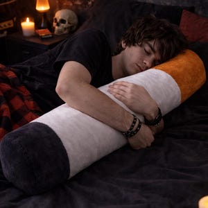 Puede incluir: Una almohada con forma de cigarrillo en blanco, naranja y negro. La almohada es cilíndrica y parece suave, descansando sobre una cama gris oscura. La imagen tiene un ambiente de dormitorio con una persona abrazando la almohada.