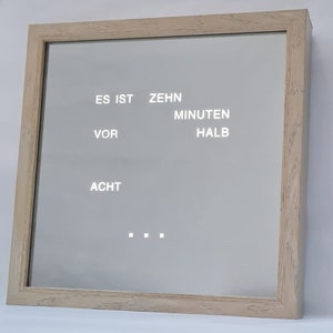 Könnte beinhalten: Ein gerahmter Spiegel mit dem Text "ES IST ZEHN MINUTEN VOR HALB ACHT" auf weißem Hintergrund.