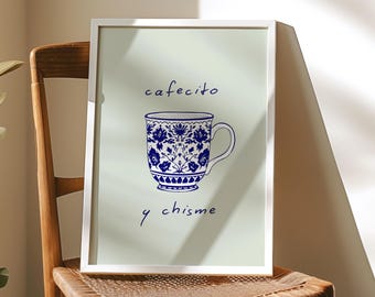 Op de afbeelding: Een ingelijste print met een blauw en wit bloemenontwerp van een koffiekopje en de woorden "cafecito y chisme" in blauw schrift. De print wordt getoond op een houten stoel met een geweven zitting.