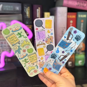 May include: Three colorful bookmarks with different designs. The first bookmark is green and yellow with a frog, a crown, and the text "There's magic in this book". The second bookmark is pink and yellow with a frying pan, a paint palette, and the text "Best Cook Ever". The third bookmark is blue and green with a bow and arrow, a book, and a bear paw.