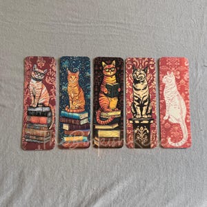 May include: Set of five colorful bookmarks featuring cats reading books. Each bookmark has a different cat design with a unique background pattern. The cats are depicted sitting on stacks of books.