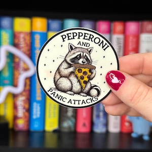 May include: A round sticker featuring a raccoon eating a slice of pizza. The sticker has the words "Pepperoni and Panic Attacks" in a circular design. The raccoon is gray and white, and the pizza slice has yellow cheese and pepperoni.