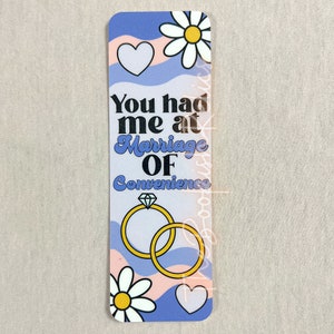 May include: A blue and pink bookmark with a white daisy, a heart, and two gold rings with a diamond. The text on the bookmark reads "You had me at Marriage of Convenience."