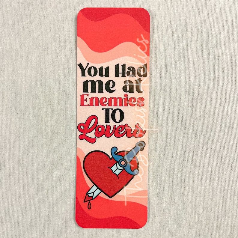 May include: A red and pink bookmark with a heart pierced by a sword and the text "You had me at Enemies to Lovers".