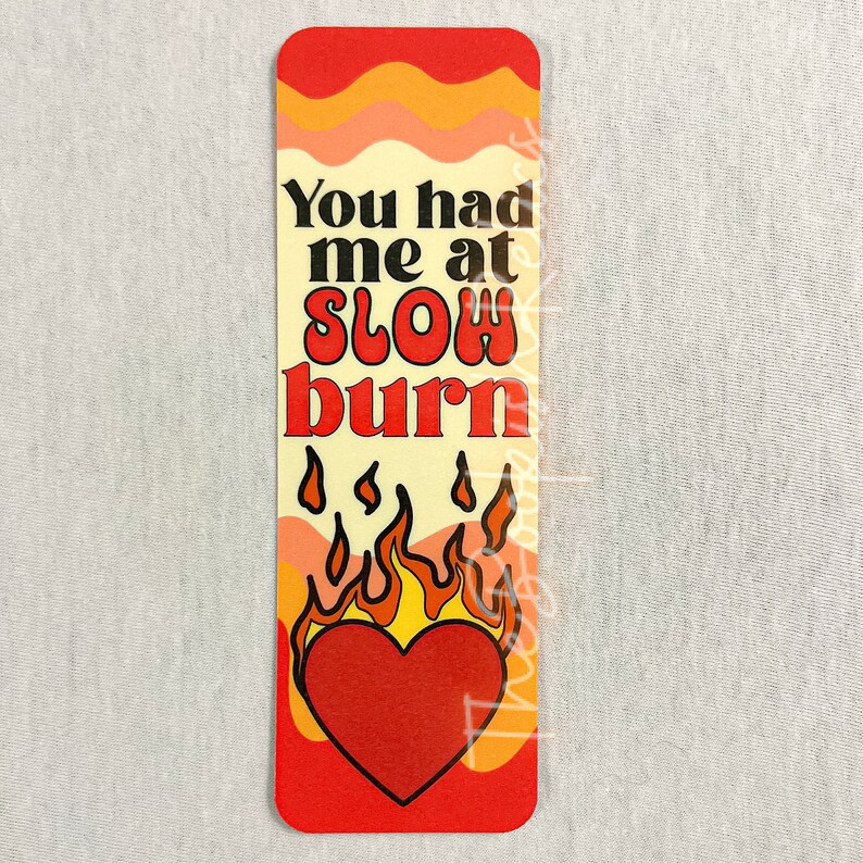 May include: A bookmark with a red heart and flames on a yellow background. The text on the bookmark reads "You had me at slow burn."