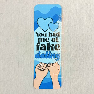 May include: Blue and white bookmark with two hearts and the text "You had me at fake dating" and two hands making a pinky promise.