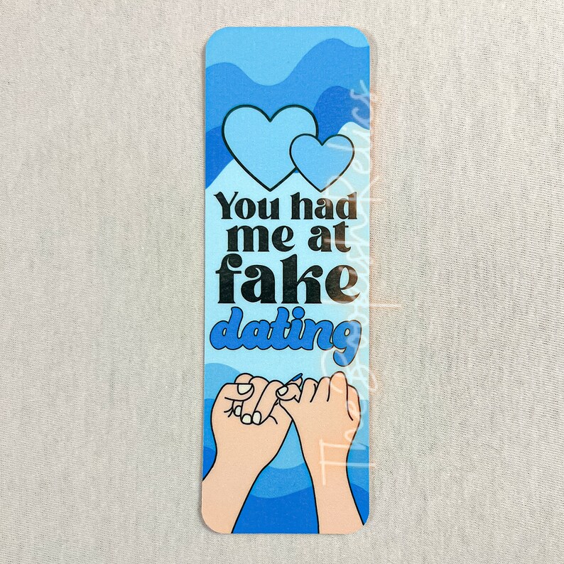 May include: Blue and white bookmark with two hearts and the text "You had me at fake dating" and two hands making a pinky promise.