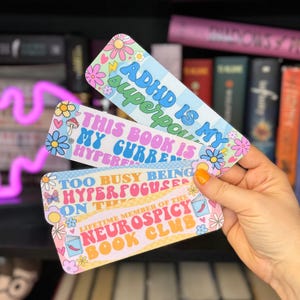 May include: Three colorful bookmarks with a retro design. The bookmarks feature text that reads "ADHD is my superpower", "This book is my current hyperfixation", and "Too busy being hyperfocused on the lifetime member of the Neurospicy Book Club".