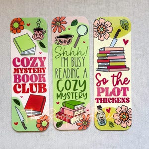 May include: Three colorful bookmarks with floral designs and text. The first bookmark says "Cozy Mystery Book Club". The second bookmark says "Shhh! I'm busy reading a Cozy Mystery". The third bookmark says "So the plot thickens".