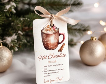 以下が含まれることがあります： ホットチョコレートのマグカップ、シナモンスティック、そして「ホットチョコレートボム」の文字が描かれた水彩画のイラストが描かれたギフトタグ。ベージュのリボンで結ばれており、説明とパーソナライズされたメッセージが記載されています。