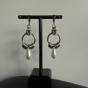 Può includere: Un paio di orecchini color argento dal design unico. Ogni orecchino presenta un grande cerchio, adornato con più anelli piccoli, e un pendente a forma di goccia di finta perla bianca. Gli orecchini sono esposti su un supporto nero.