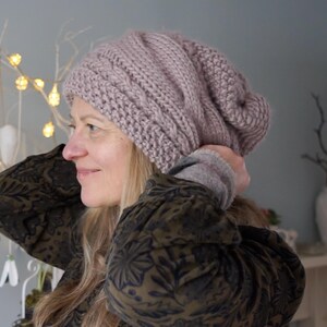 May include: A light mauve cable-knit beanie hat. The hat is made of a thick, soft yarn and has a slouchy fit. The hat is worn by a person with blonde hair and a dark green patterned jacket.