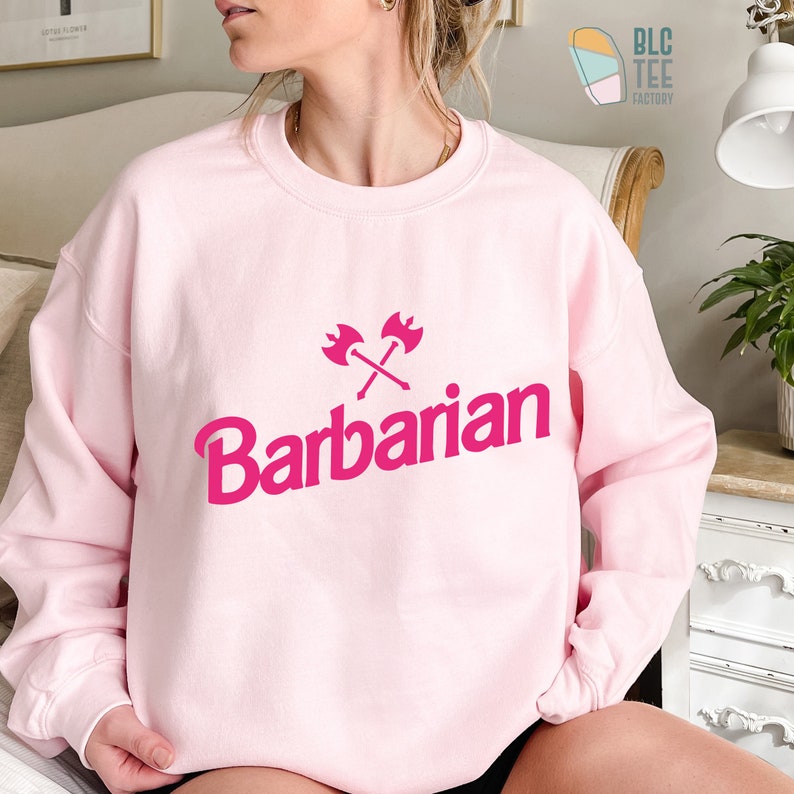 May include: Pink sweatshirt with a pink graphic of crossed axes and the word "Barbarian" in a bold, stylized font.