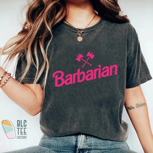 May include: A dark gray t-shirt with a pink graphic of crossed axes and the word "Barbarian".