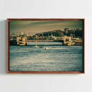 Pode incluir: Uma fotografia emoldurada de uma ponte sobre um corpo de água com barcos em primeiro plano. A ponte é feita de pedra e tem dois arcos. A água é azul escura e o céu é azul claro. O horizonte da cidade é visível no fundo.