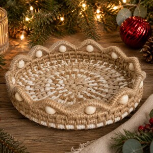 May include: A round, shallow basket crafted from natural tan and white woven cord. The rim features a scalloped edge design accented with small, smooth, round wooden beads spaced evenly along the perimeter of the woven structure.