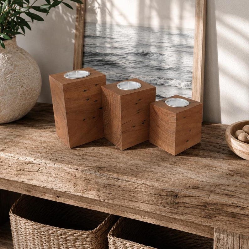May include: Three square wooden candle holders of varying heights, each featuring a smooth brown grain and a circular metal insert at the top designed to hold a tea light candle. The blocks are arranged in a cluster, showcasing natural wood textures.