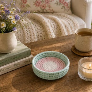 May include: A small, round, hand-woven basket featuring a light mint green exterior and a circular base with concentric rings of white and soft pink yarn. The basket has a textured, woven structure and sits on a wooden surface.