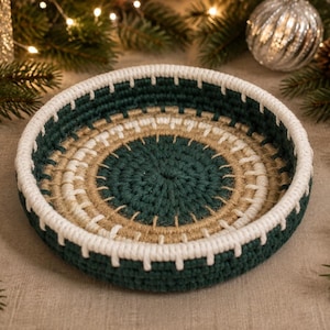 May include: A round, shallow, hand-crocheted basket featuring concentric rings of dark forest green, natural tan, and bright white yarn. The structure is firm with a thick, white, stitched rim and a flat base, showcasing a textured, woven pattern.