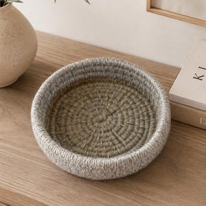 May include: A round, shallow basket constructed from coiled, textured rope. The interior base features a muted olive green hue, while the raised outer rim is crafted from a lighter, heathered grey rope, creating a distinct two-tone colour palette.