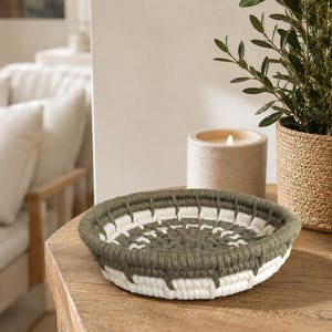 May include: A round, shallow bowl crafted from coiled rope, featuring a repeating pattern of olive green and cream white yarn wrapped around the structure. The bowl has a thick, textured rim and a flat base, showcasing a woven construction.