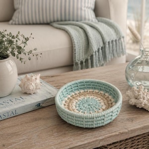 May include: A round, shallow basket featuring a concentric circular pattern in light blue, cream, and tan yarn. The basket has a thick, textured, crocheted construction with a flat base and low, vertical sides, showcasing a handmade fibre design.