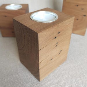 May include: A rectangular wooden candle holder crafted from natural brown timber with visible grain patterns and small dark knots. The top surface features a circular silver metal insert designed to hold a standard white tea light candle.