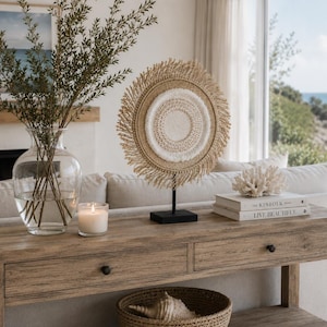 May include: A circular wall art piece with a fringed edge and a textured white centre, mounted on a black metal stand. The piece is displayed on a wooden console table alongside a clear glass vase with green branches and a white candle.