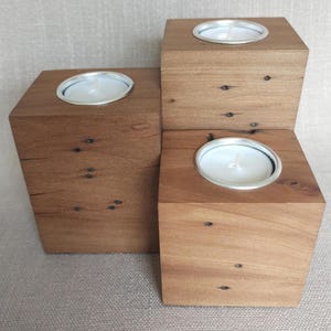 May include: Three square wooden candle holders of varying heights arranged in a cluster. Each block features a natural brown wood grain with visible knots and a circular silver metal insert at the top, holding a white tea light candle with a wick.