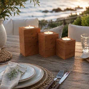 May include: Three square wooden candle holders of varying heights, each featuring a natural grain pattern and a circular top cutout holding a lit tea light candle. The holders are arranged in a cluster on a weathered wooden surface.