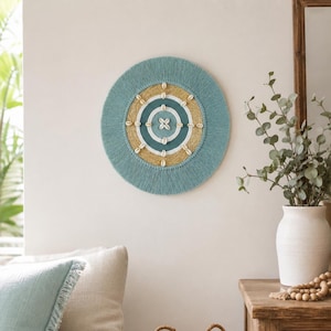 May include: A circular wall hanging featuring concentric rings of teal, white, and tan yarn. The design is accented with small white cowrie shells arranged in a radial pattern across the tan and teal sections, creating a textured, layered appearance.