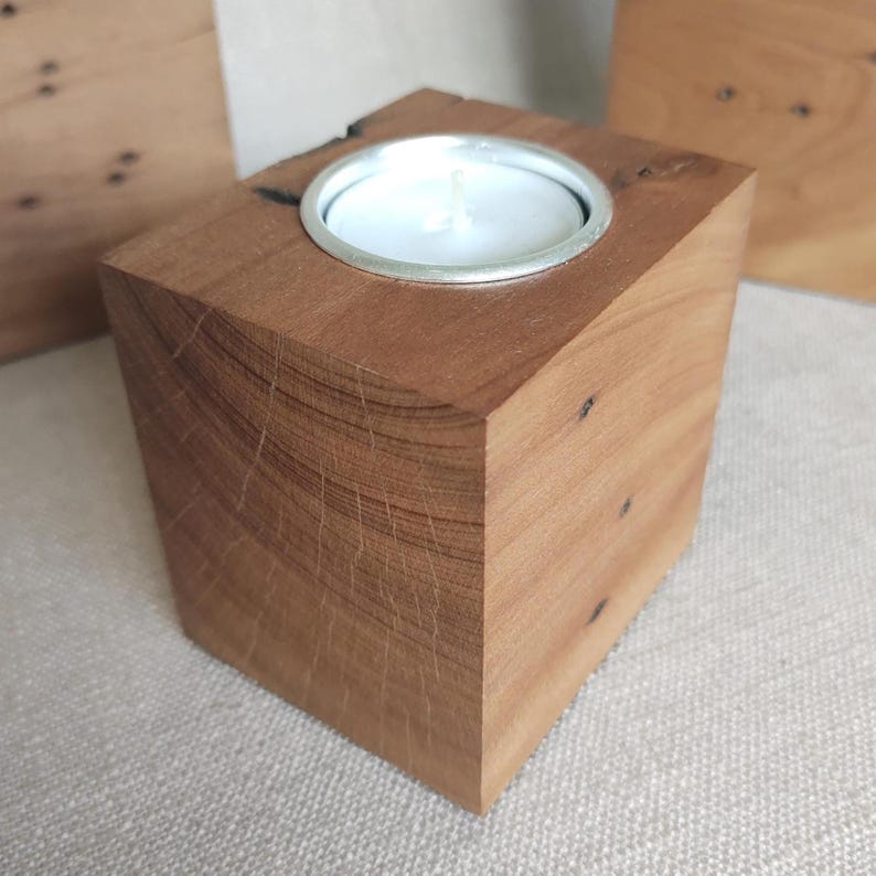 May include: A square-shaped wooden candle holder crafted from a solid block of light brown wood with visible natural grain patterns and small dark knots. A circular silver metal insert is embedded in the top center, holding a white tea light candle.