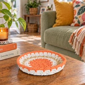 May include: A round, shallow, hand-crocheted bowl featuring concentric rings of bright orange, white, and natural tan yarn. The bowl has a thick, textured rim and a flat base, constructed with a dense stitch pattern in a circular design.
