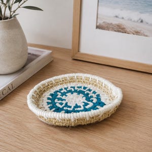 May include: A round, shallow woven tray featuring a concentric pattern of natural tan, white, and teal fibres. The tray has a raised, thick white corded rim and a flat base, constructed from coiled material with visible stitching throughout the design.