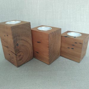 May include: Three rectangular wooden candle holders of varying heights, each crafted from natural brown timber with visible wood grain, knots, and cracks. Each block features a circular silver metal insert at the top designed to hold a tea light candle.