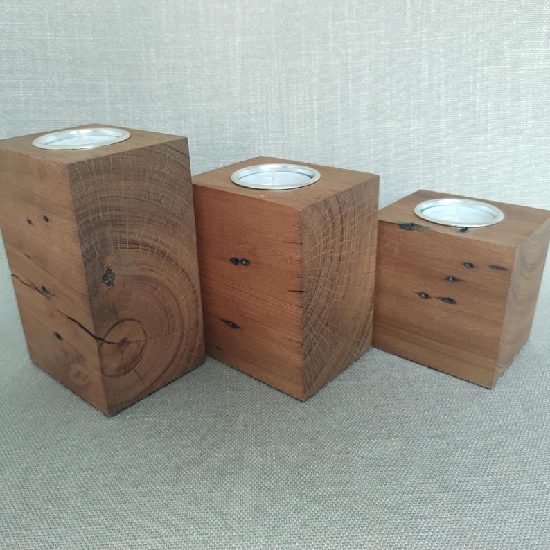 May include: Three rectangular wooden candle holders of varying heights, each crafted from natural brown timber with visible wood grain, knots, and cracks. Each block features a circular silver metal insert at the top designed to hold a tea light candle.