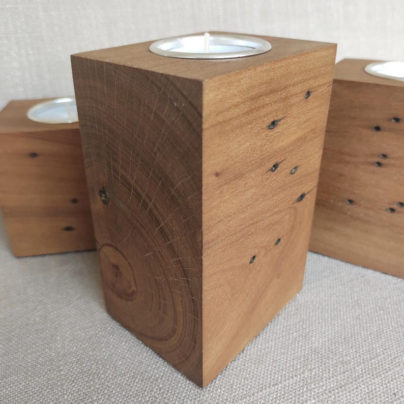 May include: A rectangular wooden candle holder featuring a natural grain pattern and visible wood knots. The top surface contains a circular metal insert designed to hold a tea light candle. The wood has a warm brown tone and a smooth finish.
