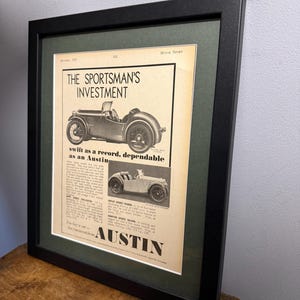 May include: A framed vintage Austin car advertisement. The black frame encloses a print featuring the headline "THE SPORTSMAN'S INVESTMENT" and illustrations of classic Austin cars. The print is set against a green mount.