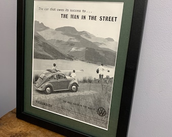 Peut inclure: Une publicité vintage en noir et blanc pour Volkswagen avec une photo d'une Volkswagen Beetle roulant sur une route avec des montagnes en arrière-plan. Le texte dit "La voiture qui doit son succès à... L'HOMME DE LA RUE LA MEILLEURE VALEUR AU MONDE VOLKSWAGEN"