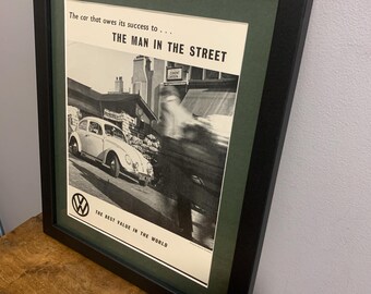 Può includere: Una pubblicità vintage in bianco e nero per un Volkswagen Maggiolino. La pubblicità presenta una foto di un Volkswagen Maggiolino bianco che guida lungo una strada. Il testo recita "L'auto che deve il suo successo a... L'UOMO DELLA STRADA" e "IL MIGLIOR VALORE AL MONDO".