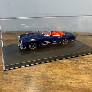 Puede incluir: Un coche modelo azul y plateado en una vitrina transparente. El coche tiene un interior rojo y es un descapotable.
