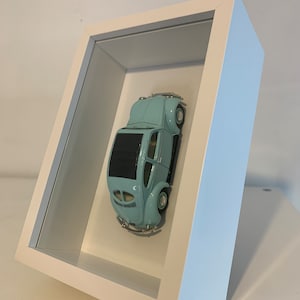 Pode incluir: Um modelo de carro vintage azul claro exibido dentro de uma moldura branca. O carro tem uma carroceria arredondada, teto preto e detalhes cromados. A moldura está inclinada, mostrando o carro em um fundo branco.