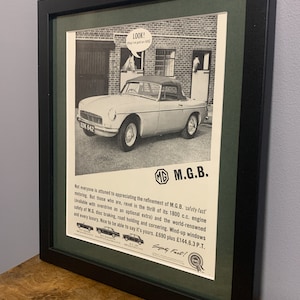 Op de afbeelding: Een zwart-witte vintage advertentie voor de M.G.B. auto. De advertentie toont een foto van een zilveren M.G.B. cabriolet die over een straat rijdt. De tekst in de advertentie luidt: "Niet iedereen is gevoelig voor de verfijning van M.G.B. 'safety-fast' rijden. Maar degenen die dat wel zijn, genieten van de sensatie van zijn 1800 cc motor (optioneel met overdrive beschikbaar) en de wereldberoemde veiligheid van M.G. schijfremmen, wegligging en bochtengedrag. Opwindbare ramen en alle luxe. Leuk om te kunnen zeggen dat hij van jou is. £690 plus £144.6.3 P.T."