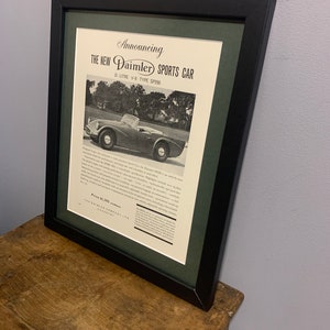 Puede incluir: Un anuncio vintage en blanco y negro para el nuevo automóvil deportivo Daimler SP250. El anuncio presenta una foto del automóvil en un entorno boscoso y un texto que dice "Anunciando El Nuevo Automóvil Deportivo Daimler 2 Litros V-8 Tipo SP250 Precio £1,395".