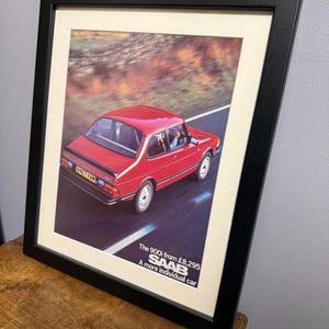 May include: Framed print featuring a red Saab 900i car speeding down a road. The image has a vintage aesthetic, with text at the bottom that reads "The 900i from £8,295 SAAB A more individual car."