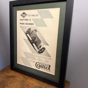 May include: A framed vintage advertisement for Riley Six-Twelve, with a black and white illustration of a race car. The ad includes the text "Shatters 3 More Records" and promotes Wakefield Castrol motor oil. The frame is black.